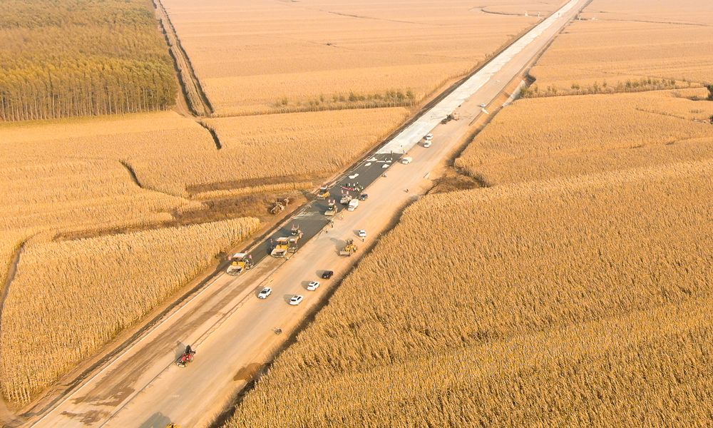 在田間地頭，農(nóng)民們忙著收玉米，將一年的勞動成果打包帶回家；在施工現(xiàn)場，建設(shè)者們揮汗如雨，全面加快路面施工。長太高速公路是吉林省中西部地區(qū)的一條重要干線通道，整條線路穿過著名的“黃金玉米帶”、我國大型商品糧和油料基地，并輻射內(nèi)蒙古東部通遼市、興安盟等多個旗縣。圖為長太高速沿路布滿玉米田。