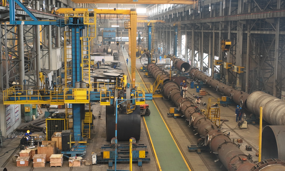 在內蒙古圖克工業區的制造現場，上百噸的“龐然大物”被拆解成一塊塊“積木”：異型錐體被分片壓制、組裝；數十噸鋼板一遍遍“翻身”，10米立車在鋼花中精雕細琢……與此同時，1500多公里外的南京車間內，工人們為巨大的“滾筒”噴上白漆，設備內焊花飛濺。這是中建安裝旗下中建五洲“現場+工廠”兩線制造模式在寶豐項目的再度應用。憑借著全國領先的大型塔器、反應器現場制造水平，項目團隊完成了內蒙古寶豐項目反應、換熱、分離及儲存容器等91臺重要設備制造與安裝。