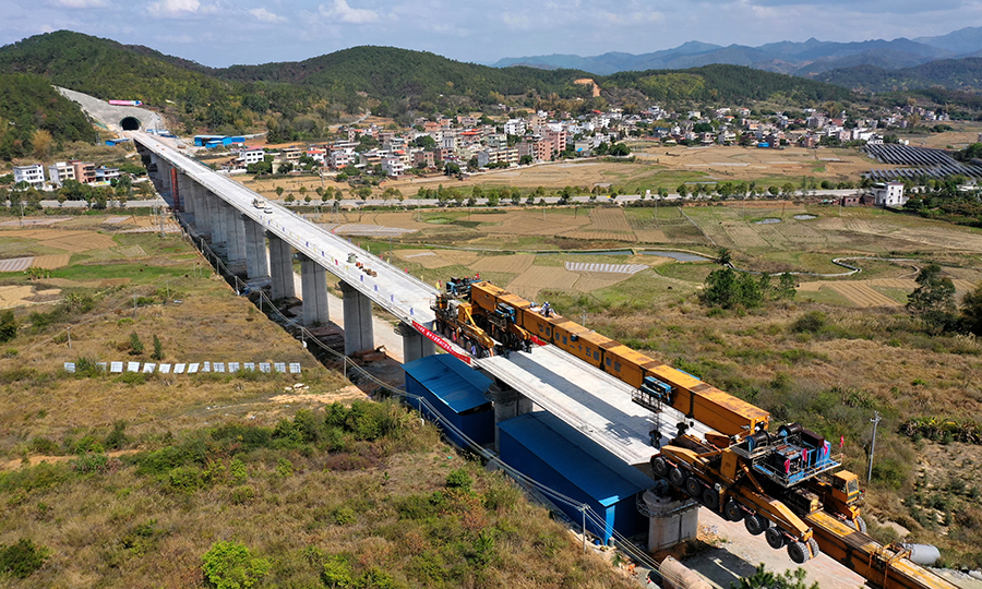 近日，國產WE-SC900型運架一體式架橋機前方伸縮支架不斷延伸，一榀820噸重、32米長的預制箱梁慢慢從尾部移動到“腹中”，穩穩落在贛深鐵路佳派跨G205國道特大橋的橋墩之間。粵港澳大灣區重點項目贛深鐵路項目架梁施工有序推進。圖為架梁施工俯瞰。（陳浩 攝）